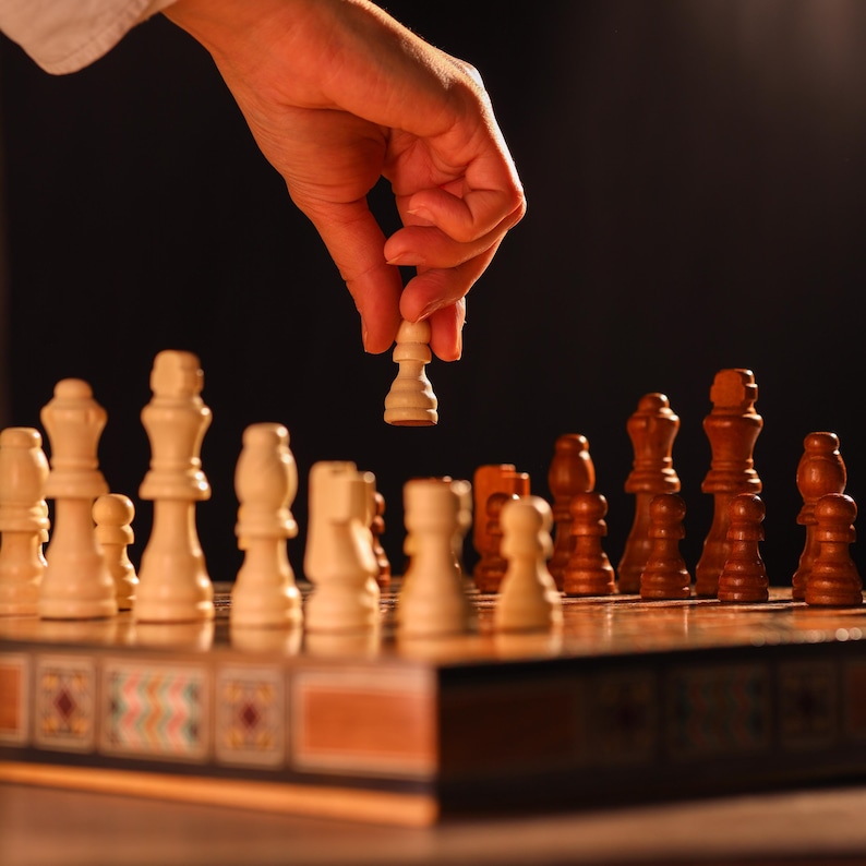 May include: A hand reaches down to move a white chess piece on a wooden chessboard. The chessboard has a decorative border and is filled with white and brown chess pieces.
