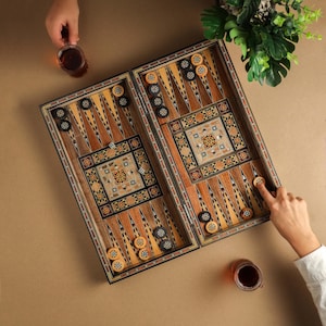 May include: A wooden backgammon game board with intricate inlaid designs. The board is open and shows black and white game pieces, as well as a few brown game pieces. Two glasses of tea are visible on the table.