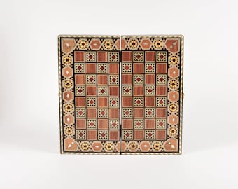Op de afbeelding: Een opvouwbaar houten spelbord met een ingewikkeld inlegwerkontwerp. Het oppervlak toont een raster van afwisselend lichte en donkere houten vierkanten, omringd door een decoratieve rand van geometrische sterpatronen, ruiten en ingelegd parelmoer.