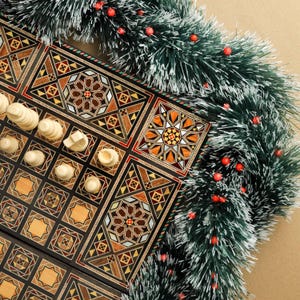 May include: A wooden chessboard with intricate inlaid designs in shades of brown, black, and white. The board is partially covered with white chess pieces, and a green and white garland with red berries is draped over the top.