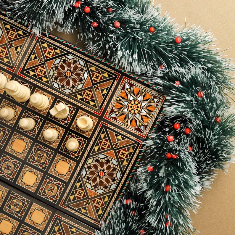 May include: A wooden chessboard with intricate inlaid designs in shades of brown, black, and white. The board is partially covered with white chess pieces, and a green and white garland with red berries is draped over the top.
