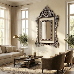May include: An ornate, dark-framed mirror with a detailed, carved design hangs on a cream-colored wall. The mirror is the focal point of a living room, complemented by neutral-toned furniture, a coffee table, and a patterned rug.