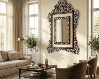 May include: An ornate, dark-framed mirror with a detailed, carved design hangs on a cream-colored wall. The mirror is the focal point of a living room, complemented by neutral-toned furniture, a coffee table, and a patterned rug.