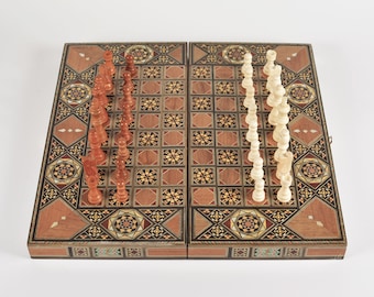 Peut inclure: Un échiquier en bois avec des motifs incrustés complexes. Le plateau est ouvert et montre un jeu complet de pièces d'échecs, avec les pièces blanches à droite et les pièces brunes à gauche.
