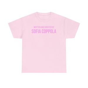Puede incluir: Una camiseta rosa claro con el texto "WRITTEN AND DIRECTED BY SOFIA COPPOLA" en rosa a juego. La camiseta es de cuello redondo.
