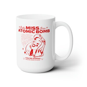 May include: White ceramic mug with a red graphic design featuring a woman with the text "Battle Miss Born Atomic Bomb Burnin' for Love You're Gonna Miss Me When I'm Gone"