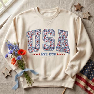 Könnte beinhalten: Ein cremefarbenes Sweatshirt mit Rundhalsausschnitt und den Buchstaben USA in einer blau umrandeten Blockschrift, die mit einem rot-weiß-blauen Blumenmuster gefüllt ist. Unter den Buchstaben steht der Text EST. 1776, flankiert von kleinen roten Sternen.