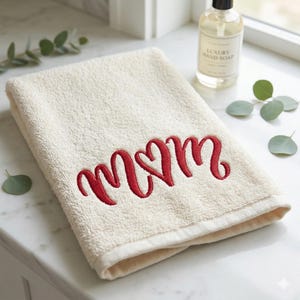 Puede incluir: Toalla plegada de color crema con la palabra "MOM" bordada en rojo. La toalla está hecha de un material suave y absorbente, probablemente un accesorio de baño o cocina. Una botella de jabón de manos está en el fondo.