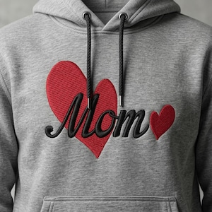 Puede incluir: Sudadera con capucha gris con un corazón rojo y la palabra "Mom" bordada en escritura negra. Un corazón rojo más pequeño está a la derecha de la palabra. La sudadera tiene un bolsillo delantero y cordones negros.
