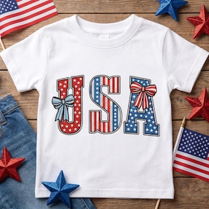Könnte beinhalten: Ein weißes Kurzarm-T-Shirt mit dem Schriftzug USA in einem patriotischen Design. Das U ist rot mit weißen Sternen und einer blauen Schleife, das S hat rote und weiße Streifen und das A ist blau mit weißen Sternen und einer rot-weiß gestreiften Schleife.