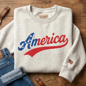 Peut inclure: Un sweat-shirt à col rond gris clair chiné avec le mot America brodé en lettres cursives bleues et rouges sur la poitrine. La lettre A comporte des étoiles blanches. Un petit écusson drapeau américain est sur le poignet gauche.