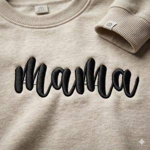 Op de afbeelding: Een beige sweatshirt met het woord "Mama" in zwarte cursieve letters op de voorkant geborduurd. De sweatshirt heeft een ronde hals en geribde manchetten. De manchet van de mouw is zichtbaar in de afbeelding.