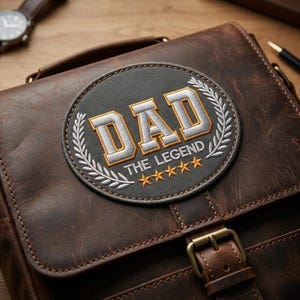 Puede incluir: Bolso mensajero de cuero marrón con un parche circular que dice "DAD THE LEGEND" en letras blancas y doradas. El parche está rodeado por una corona de laurel plateada y cinco estrellas doradas. El bolso tiene un cierre de hebilla.