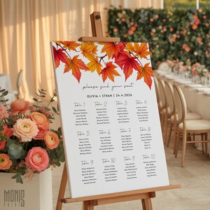 May include: A white wedding seating chart sign on a wooden easel, decorated with red and orange maple leaves at the top. The text reads "please find your seat", "OLIVIA + ETHAN | 24.4.2026", and lists guest names under twelve numbered tables.