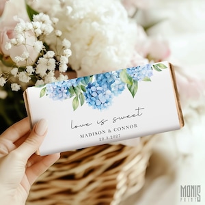 以下が含まれることがあります： 青いアジサイの花と緑の葉の水彩画が描かれた長方形のチョコレートバーの包装紙。白いラベルには、黒い文字で「love is sweet」、「MADISON & CONNOR」、日付「22.3.2027」が印刷されています。