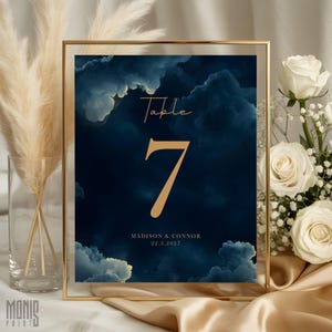 May include: A rectangular table number sign in a thin gold frame featuring a dark navy blue background with a cloud pattern. The sign displays the text "Table 7" in gold, followed by the names "MADISON & CONNOR" and the date "22.3.2027" in gold.