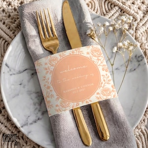 May include: A gold fork and knife wrapped in a grey napkin with a peach-colored paper band featuring floral illustrations and the text: welcome to the wedding of MADISON & CONNOR 22.3.2027, placed on a white marble plate with a sprig of baby's breath.