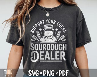 Puede incluir: Una camiseta gris carbón de manga corta con un estampado gráfico blanco de un frasco y una hogaza de pan, junto al texto "SUPPORT YOUR LOCAL SOURDOUGH DEALER" dispuesto en un diseño circular y en bloque con espigas y estrellas.