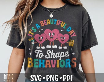 May include: A charcoal gray t-shirt featuring a colorful graphic of three pink heart characters with faces, arms, and legs. The text reads IT'S A BEAUTIFUL DAY To Shape BEHAVIORS. One heart holds yellow flowers, another holds a gift box, and stars surround them.