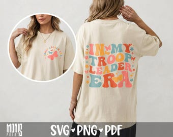 Könnte beinhalten: Ein cremefarbenes T-Shirt mit Grafikdruck. Auf der Rückseite steht der Text IN MY TROOP LEADER ERA in großen, bunten, wellenförmigen Buchstaben, umgeben von kleinen Schmetterlingen und Blumen. Die Vorderseite zeigt ein kleineres Motiv.