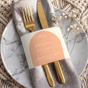 May include: A gold fork and knife wrapped in a grey linen napkin with a peach-colored paper band featuring the text M|C, welcome to the wedding of, MADISON & CONNOR, and 22.3.2027, placed on a white marble plate with a sprig of white baby's breath flowers.