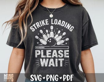 May include: A charcoal gray short-sleeve t-shirt featuring a white graphic print of bowling pins and a bowling ball. The text reads STRIKE LOADING PLEASE WAIT above and below the graphic, which includes a loading bar design element.