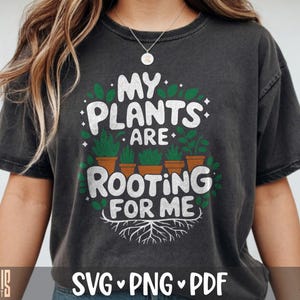 May include: A charcoal gray short-sleeve t-shirt featuring a graphic print with the text "MY PLANTS ARE ROOTING FOR ME" in white letters. The design includes five potted green plants and white root illustrations at the bottom of the text.