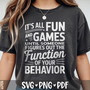 Puede incluir: Una camiseta de manga corta en color gris carbón con texto blanco que dice: IT'S ALL FUN AND GAMES UNTIL SOMEONE FIGURES OUT THE FUNCTION OF YOUR BEHAVIOR. La prenda tiene cuello redondo y un aspecto de tela lavada y desgastada.