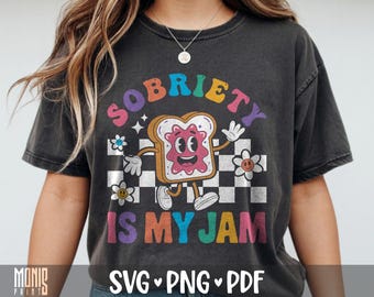 以下が含まれることがあります： チャコールグレーのTシャツで、笑顔のトーストのイラストとピンクのジャムが描かれています。SOBRIETY IS MY JAMという文字がカラフルなフォントで書かれ、白黒のチェック柄と小さな花々が周囲を囲んでいます。
