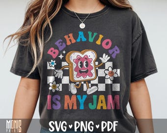 May include: A charcoal gray t-shirt featuring a graphic of a cartoon slice of toast with pink jam, arms, and legs. The text reads BEHAVIOR IS MY JAM in colorful letters. The design includes a black and white checkered background and small flowers.