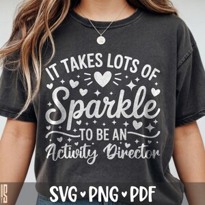 May include: A charcoal gray short-sleeve t-shirt featuring white text that reads IT TAKES LOTS OF Sparkle TO BE AN Activity Director, surrounded by various heart and star icons. The design is centered on the front of the garment.