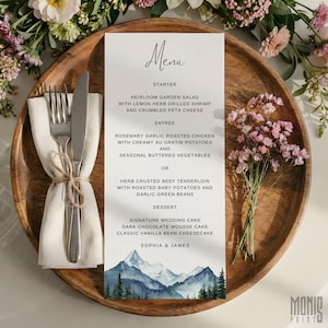 Könnte beinhalten: Eine Hochzeitsmenükarte mit einem Aquarell-Bergdesign, platziert auf einem Holz-Ladegerät mit Besteck und einer Serviette. Die Speisekarte enthält Vorspeisen-, Hauptspeisen- und Dessertoptionen. Blumengestecke umgeben den Teller.