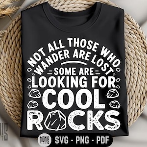 Może przedstawiać: Czarny T-shirt z białym tekstem, który brzmi "Not all those who wander are lost - some are looking for cool rocks". Tekst jest otoczony małymi białymi kształtami skał.