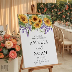 Könnte beinhalten: Ein Hochzeits-Willkommensschild auf einer Holzstaffelei. Das Schild zeigt ein Blumendesign aus Sonnenblumen und Lavendel. Der Text lautet "Willkommen zu unserer Hochzeit" mit den Namen "Amelia und Noah" und dem Datum 29.11.2026. Das Schild sagt auch "so glad you're here!"