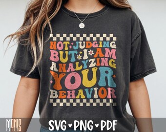 May include: A charcoal gray t-shirt featuring a graphic print with the text NOT JUDGING BUT I AM ANALYZING YOUR BEHAVIOR in colorful, wavy, retro-style lettering. The design is framed by a checkered border at the top and bottom of the text.