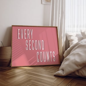 May include: A pink framed poster with the text "EVERY SECOND COUNTS" in white letters. The poster is hanging on a white wall in a room with a bed and a window.