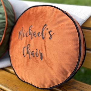 Puede incluir: Un cojín redondo de terciopelo naranja con ribete negro y el texto "Michael's Chair" bordado en negro.