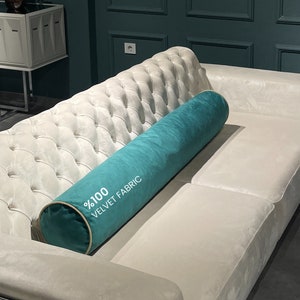 Könnte beinhalten: Ein weißes, gestepptes Sofa mit einem zylindrischen Kissen aus türkisfarbenem Samt, auf dem "100% Samtstoff" steht.