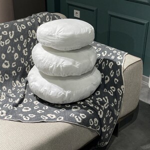May include: Three white round cushions stacked on top of each other on a gray and white patterned throw blanket. The cushions are made of a soft, plush fabric.