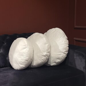 May include: Three white round pillows of different sizes are arranged on a black velvet sofa. The pillows are made of a soft, plush fabric and have a simple, elegant design.