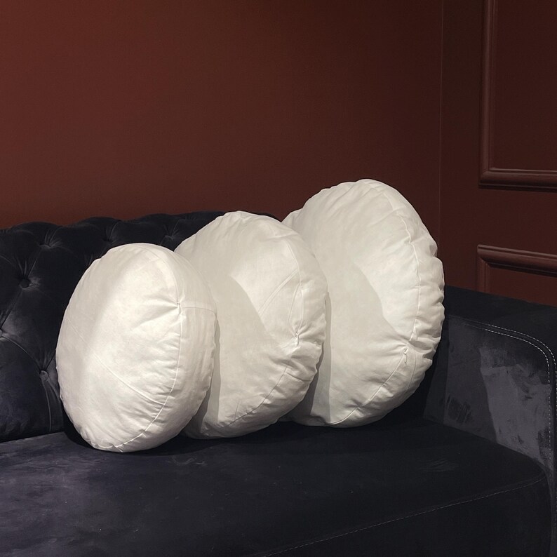 May include: Three white round pillows of different sizes are arranged on a black velvet sofa. The pillows are made of a soft, plush fabric and have a simple, elegant design.