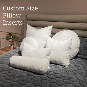 May include: A variety of white pillow inserts of different shapes and sizes, including a round pillow, a rectangular pillow, and a bolster pillow. The pillows are arranged on a gray quilted bedspread. The text "Custom Size Pillow Inserts" is displayed at the top of the image.