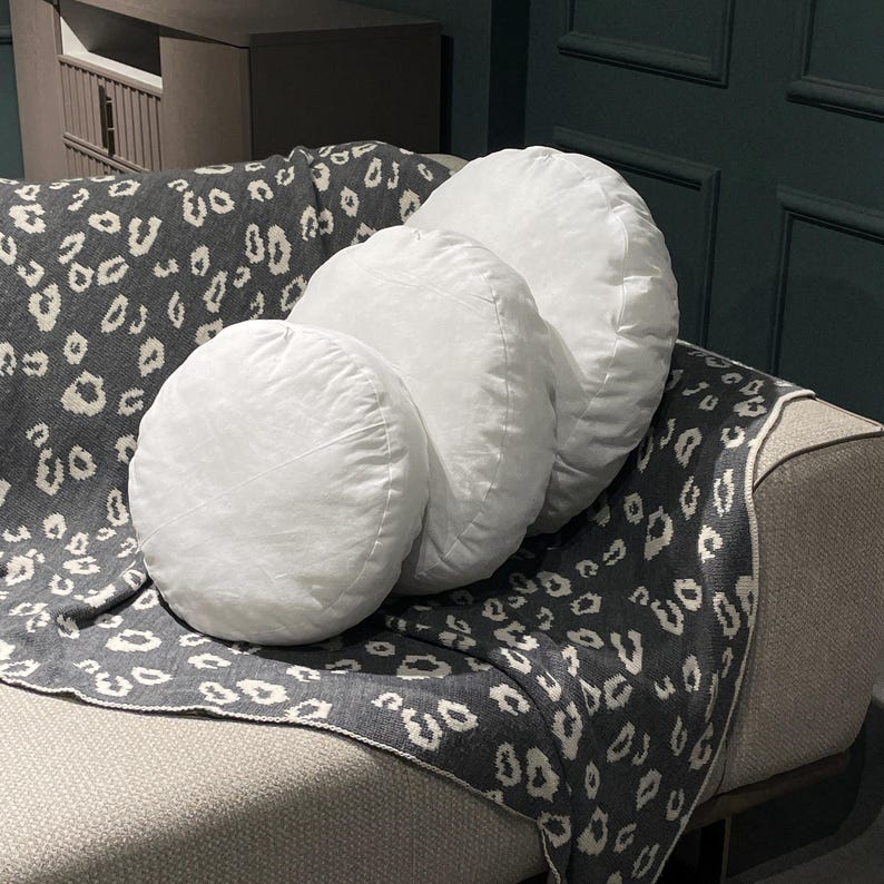 May include: Four white round throw pillows with a soft, plush texture are arranged on a gray and white patterned throw blanket. The pillows are arranged in a staggered formation, with the largest pillow at the back and the smallest pillow at the front.