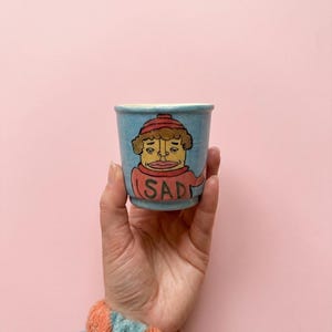 May include: A small, light blue ceramic cup with a cartoon illustration of a sad-looking face wearing a red hat and sweater. The word "SAD" is written on the cup. The cup is held against a pink background.
