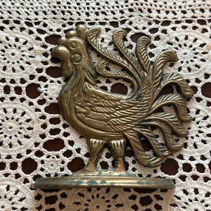 May include: A brass rooster figurine with detailed feathering and a decorative base. The rooster is standing on a small, round base. The figurine has a vintage look with some green patina.
