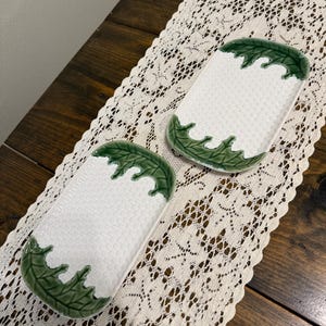 May include: Two rectangular white ceramic serving dishes with a textured surface. Each dish has a green leaf design around the edges. The dishes are displayed on a cream-colored lace tablecloth.