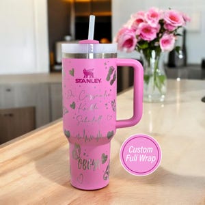 May include: Pink Stanley tumbler with a white lid and handle. The tumbler features custom silver engravings of hearts, butterflies, footprints, and text. A straw is inserted into the lid. The text on the tumbler reads "Dr. Cassandra Kuetta Scheffelt" and "OBGYN".