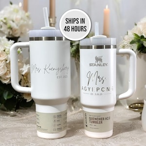 May include: Two white stainless steel travel mugs with handles and lids. The mugs are personalized with the text "Mrs. Koenigsbery EST. 2025" and "Mrs. Agyepong EST. 5.31.25". The mugs are both labeled "Stanley" and "The Flowstate Quencher H2.0 Tumbler".