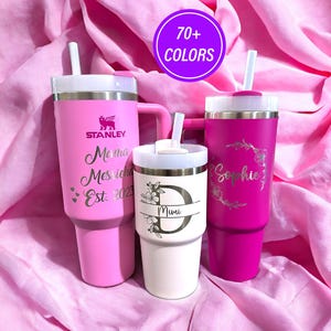 May include: Three stainless steel tumblers with lids and straws. The tumblers are pink, white, and a darker shade of pink. The tumblers are personalized with the names "Mama Messiche Est. 2023", "Mimi", and "Sophie".