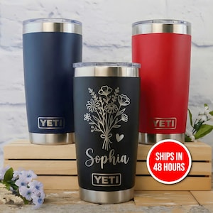May include: Three insulated tumblers in navy blue, black, and red. The black tumbler features a floral design and the name "Sophia." Each tumbler has a silver base and the "YETI" logo. The tumblers are displayed on a wooden box.